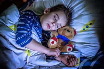 En gutt ligger og sover med en kosebamse inntil seg. 