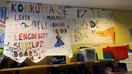 plakater skrevet av barn henger på en vegg, de handler om ulike leker 