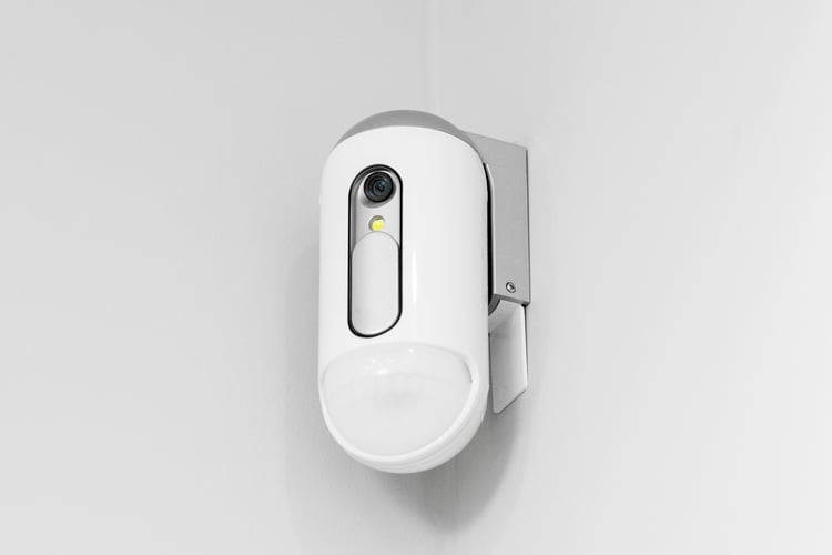 Et hvitt overvåkningskamera henger på en hvit vegg. 