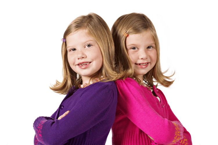 To tvillinger står med armene i kors og ryggen mot hverandre, mens de smiler mot kameraet.