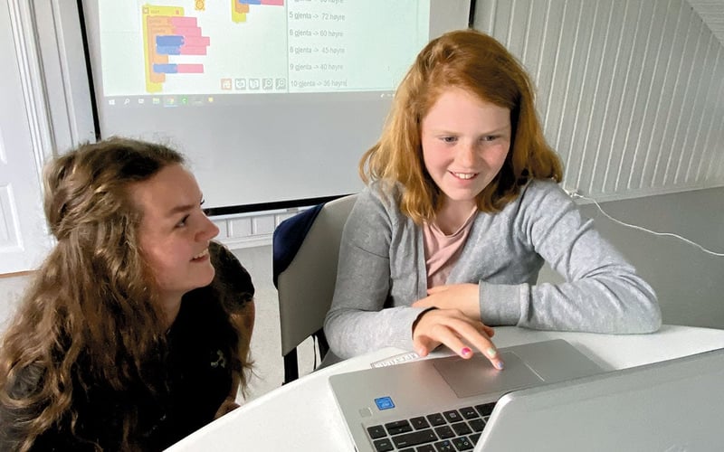 Jente får hjelp av kvinne ved datamaskin med kodeprogram i bakgrunnne