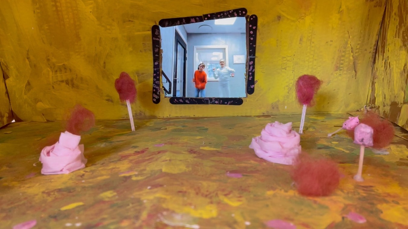 Inni den glade boksen er veggene gule, det er røde og rosa blomster av papir og vatt rundt i boksen og på bakveggen er det en video av barn som danser og synger