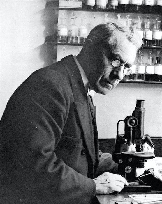 Gammelt svarthvitt foto av mann med mikroskop og små glassflasker i bakgrunnen